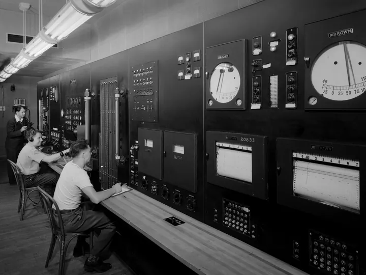 Photo d’origine : Salle de contrôle d'une soufflerie : mur de cadrans, de voyants et d'enregistreurs à papier, surveillé par des opérateurs.