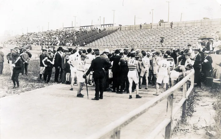 Photo d’origine : Des coureurs en tenue d'époque, dossards sur le dos, rassemblés sur la ligne de départ d'un stade devant les tribunes.