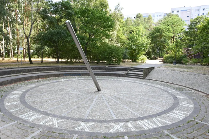 Original photo: Un grand cadran solaire au sol dans un parc, avec son style métallique incliné et ses heures en chiffres romains.