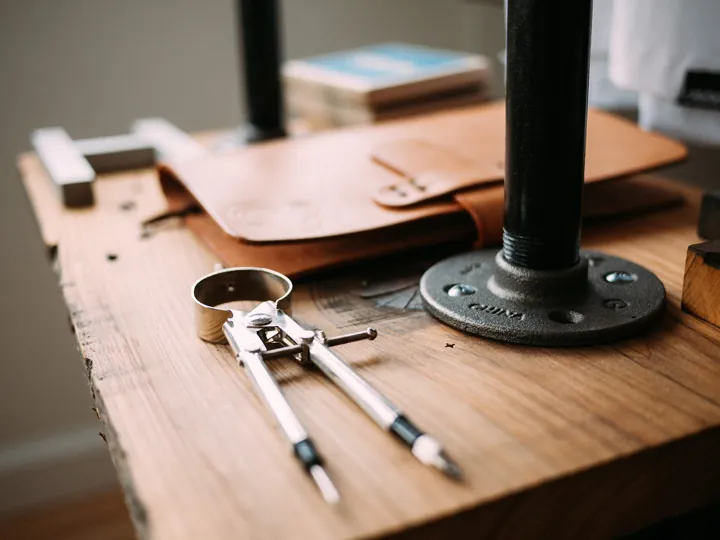 Photo d’origine : Un établi en bois avec un compas à pointes sèches, un carnet de cuir et un pied de lampe en tuyau.